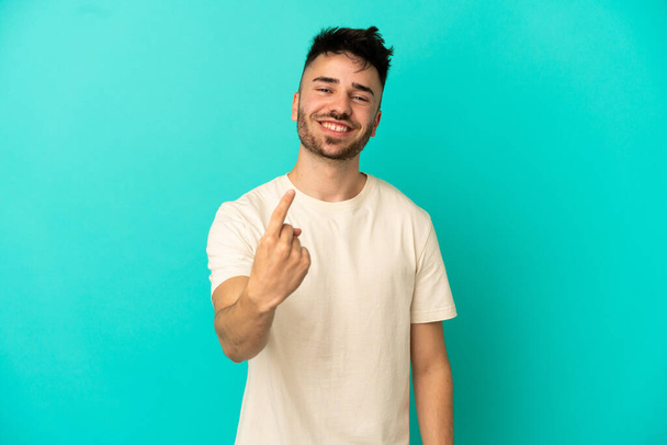 Young caucasian man isolated on blue background doing coming gesture - Photo, Image