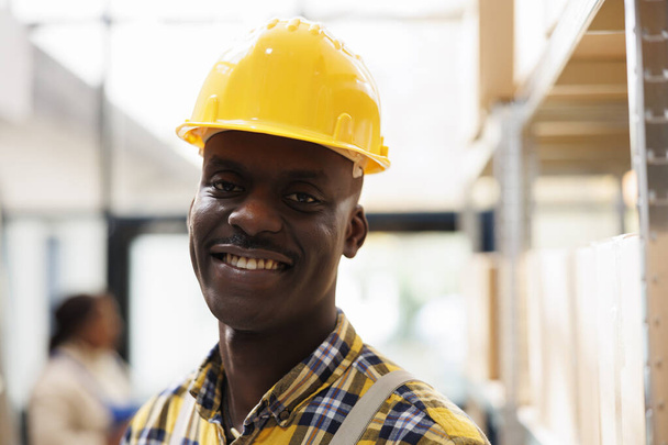Африкансько-американський обробник пакунків у жовтому шоломі. Промисловий склад молодого навантажувача в захисному одязі посміхається і дивиться на камеру крупним планом - Фото, зображення