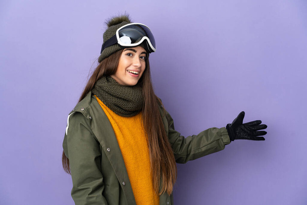 Скромна біла дівчина зі сноубордистськими окулярами ізольована на фіолетовому фоні, що розтягує руки на бік, щоб запросити прийти
 - Фото, зображення