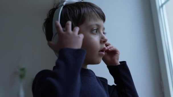 One small boy wearing headphones standing by window, child takes off headset figuring out how device works. Kid listening to music, song, audio - Footage, Video