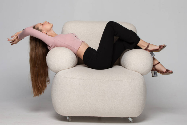 Una hermosa chica delgada con el pelo largo y rubio se encuentra en una cómoda silla blanca. Una mujer descansa y se relaja en una silla. - Foto, Imagen