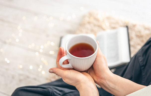 Une tasse de thé dans les mains des femmes gros plan. Une femme lit un livre assis sur un tapis par terre dans une pièce, copie l'espace. - Photo, image