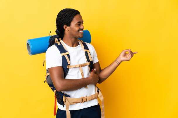 Joven montañero con trenzas con una gran mochila aislada sobre fondo amarillo apuntando con el dedo hacia un lado - Foto, imagen