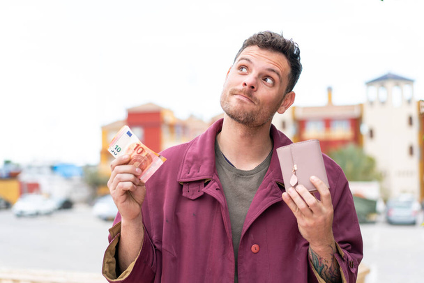 Joven hombre caucásico al aire libre sosteniendo la billetera con dinero y buscándola - Foto, imagen