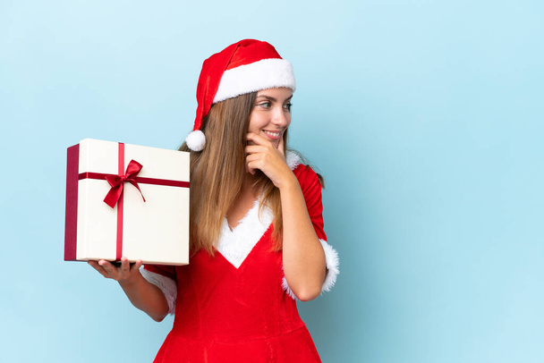 Anne Noel gibi giyinmiş genç beyaz bir kadın elinde hediyeyle mavi arka planda izole edilmiş bir şekilde bir fikir ve bir taraf arıyor. - Fotoğraf, Görsel