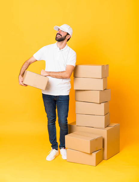 Full-length tiro de homem de entrega entre caixas sobre fundo amarelo isolado feliz e sorrindo - Foto, Imagem