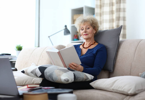 Elderly woman is reading book at home on couch. Home leisure pensioners concept - Zdjęcie, obraz