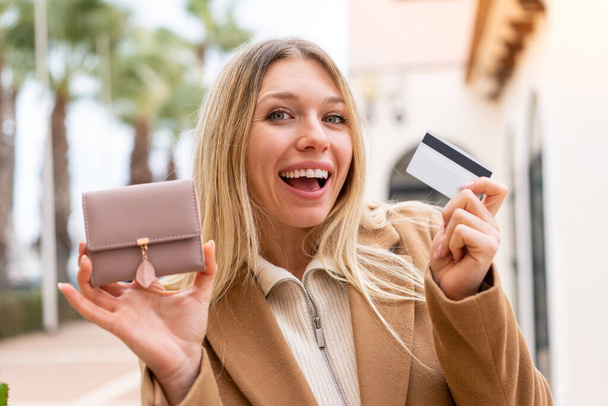 Jonge mooie blonde vrouw met portemonnee en het houden van een creditcard in de buitenlucht - Foto, afbeelding