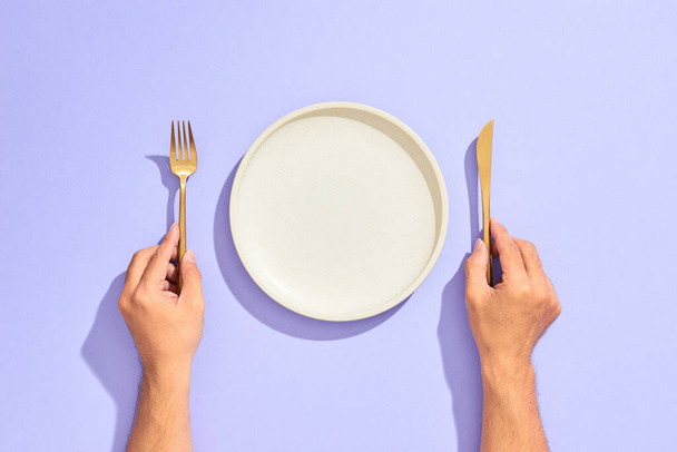 Βραδινό σκηνικό. Λευκό άδειο πιάτο και χέρι κρατώντας το χρυσό πιρούνι και μαχαίρι σε λιλά φόντο - Φωτογραφία, εικόνα
