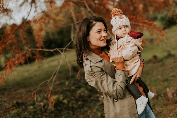 秋の公園でかわいい赤ん坊の女の子を抱えている若い女性 - 写真・画像