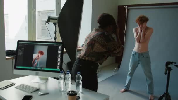 Nuori paidaton miesmalli sinisissä farkuissa poseeraa harmaalla cyc seinällä studiossa, kun taas naisvalokuvaaja ottaa kuvia muotikuvausten aikana - Materiaali, video