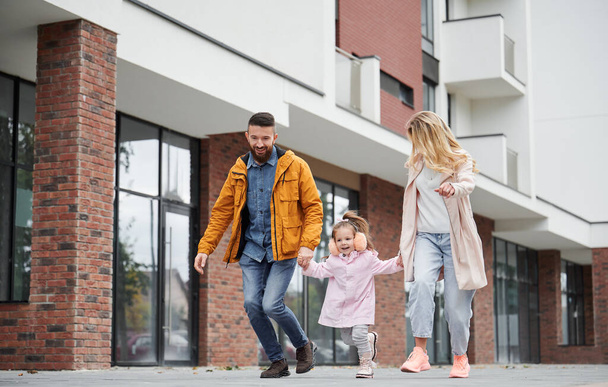 Full length of man and woman holding hands of little girl while walking down the street in new urban district. Happy loving family with child enjoying stroll outdoors near residential building. - Foto, imagen