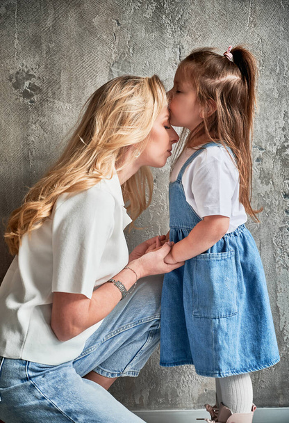 Side näkymä söpö lapsi suudella äiti seisoessaan seinää vasten kotona. Blondi nainen pitää tyttärensä kädestä, kun lapsi asettaa suudella äidin otsaan. Perhe ja äitiys käsite. - Valokuva, kuva