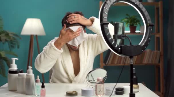 Hombre blogger con un teléfono móvil está grabando un vlog de belleza, filmando diariamente tutorial de rutina de cuidado de la piel en la cámara, cara de limpiador con servilleta. Copiar espacio - Imágenes, Vídeo