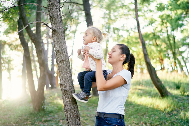Mama podnosi małą dziewczynkę na wyciągniętych ramionach na drzewo w parku. Wysokiej jakości zdjęcie - Zdjęcie, obraz