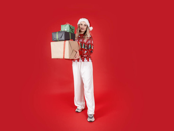 Trzymając stos pudełek z prezentami, portret młodej Mikołajki trzymającej stos pudełek z prezentami. Świętujemy święta Bożego Narodzenia. Zakupy dają prezenty. Pojedyncze czerwone tło.  - Zdjęcie, obraz