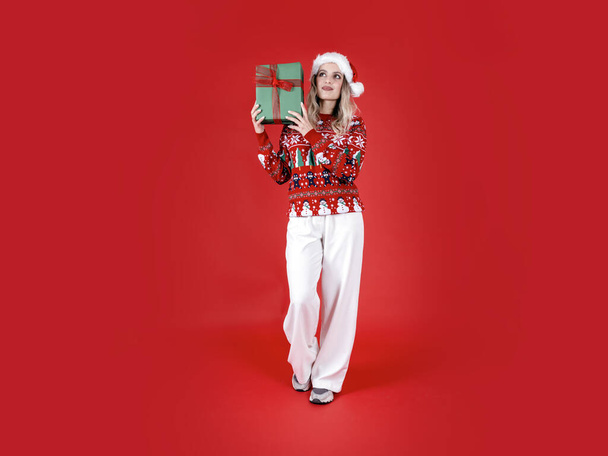 Тримає різдвяну подарункову коробку, повне тіло Санта-Жінка тримає різдвяну подарункову коробку. Щаслива кавказька блондинка бере подарункову коробку, тремтячи. Одягни Санта-капелюх, светр. Червоний фон, простір копіювання.  - Фото, зображення