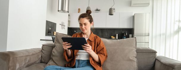 Portré fiatal nő ül a kanapén a nappaliban, filmeket néz digitális táblagépen, olvasás készüléken, élvezi a hétvégét otthon. - Fotó, kép