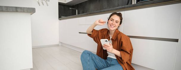 Femme moderne souriante à la maison, assise sur le sol de la cuisine avec téléphone portable, portant un casque noir sans fil, écoutant de la musique ou un podcast, regardant des vidéos sur l'application smartphone. - Photo, image