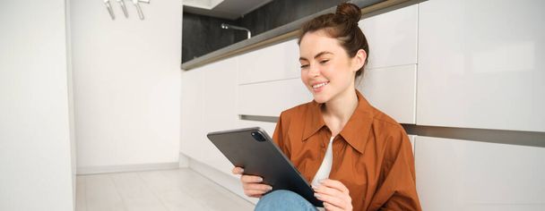 Πορτρέτο της όμορφης νεαρής γυναίκας στο σπίτι, κάθεται στο πάτωμα της κουζίνας, κοιτάζοντας ψηφιακή ταμπλέτα, χρησιμοποιώντας gadget, ανάγνωση στη συσκευή. - Φωτογραφία, εικόνα