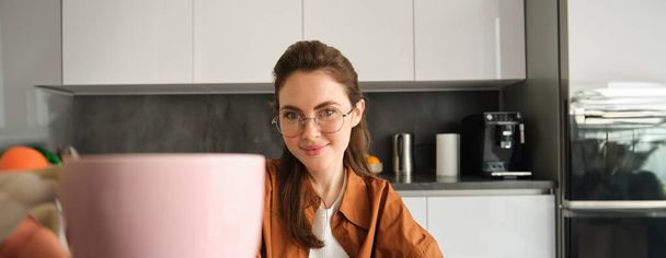 Close up portrait of beautiful young woman, giving you cup of coffee, inviting for tea, sitting in kitchen and smiling. - Valokuva, kuva