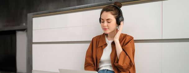 Mujer joven, estudiante haciendo los deberes en el portátil, sentado en el suelo, escuchando música en los auriculares y trabajando desde casa. - Foto, imagen