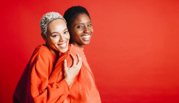 Portrait of two young female couple, wearing red sweater, embracing in front of a red background at studio. Two women together at studio. - Photo, Image