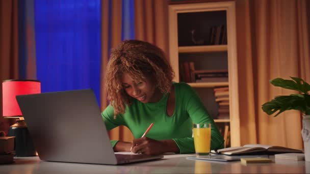Молодая африканская студентка использует ноутбук для поиска необходимой информации для подготовки к экзамену и пишет в блокноте, используя левую руку. Женщина учит иностранный язык, смотрит на монитор - Кадры, видео