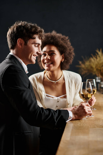 χαρούμενη Αφροαμερικανή γυναίκα που κρατάει ποτήρι κρασιού και κοιτάζει τον άντρα κατά τη διάρκεια του ραντεβού την ημέρα του Αγίου Βαλεντίνου - Φωτογραφία, εικόνα