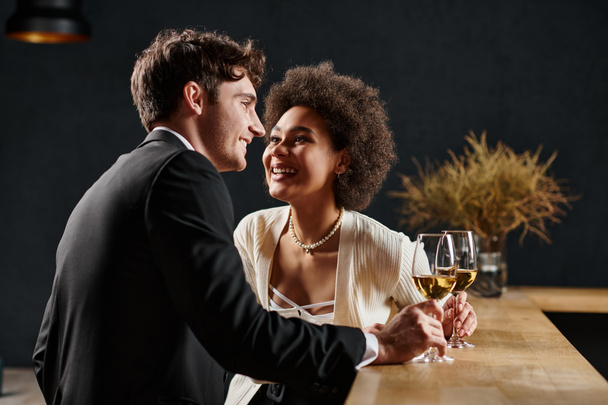 χαρούμενη Αφροαμερικανή κυρία που κρατάει ποτήρι κρασιού και κοιτάζει τον άντρα κατά τη διάρκεια του ραντεβού την ημέρα του Αγίου Βαλεντίνου - Φωτογραφία, εικόνα