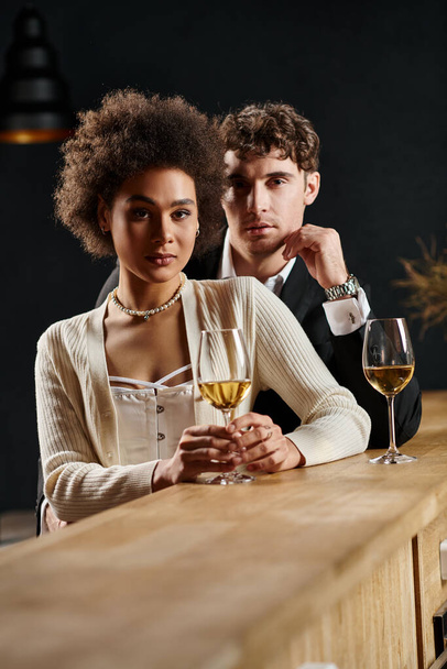 ελκυστικό πολυπολιτισμικό ζευγάρι κοιτάζοντας κάμερα, ενώ στέκεται κοντά μπαρ μετρητή με ποτήρια κρασιού - Φωτογραφία, εικόνα