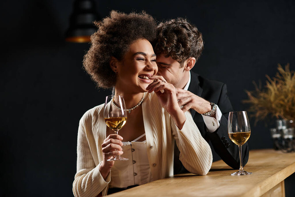 happy multicultural couple laughing while sitting at bar counter with wine glasses during date - Photo, Image