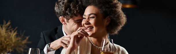 happy multicultural couple laughing and sitting at bar counter with wine glasses during date, banner - Photo, Image