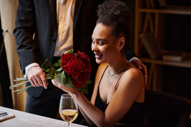 Mann im Anzug hält Rosen neben glücklicher junger afrikanisch-amerikanischer Frau in Restaurant am Valentinstag - Foto, Bild