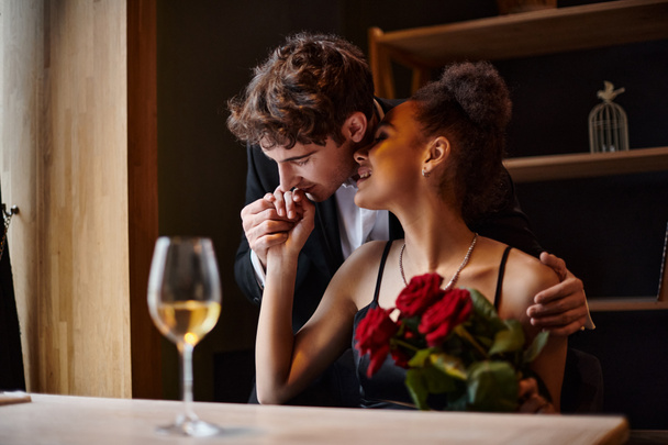 gentleman kissing hand of happy african american woman holding roses during date on Valentines day - Photo, Image