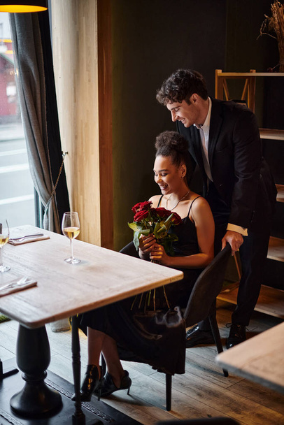 gentleman standing behind chair of happy african american woman holding roses during date - Photo, Image