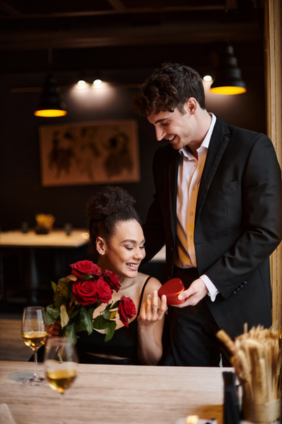 caballero regalando caja en forma de corazón a la mujer afroamericana feliz con rosas rojas el 14 de febrero - Foto, imagen