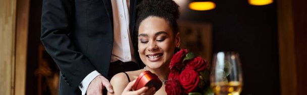 man schenkt hartvormige doos aan gelukkige Afrikaans-Amerikaanse vrouw met rode rozen op 14 februari, spandoek - Foto, afbeelding