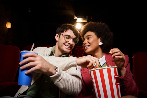 vrolijk jong multicultureel paar in retro kleding genieten van date in de bioscoop en het delen van popcorn - Foto, afbeelding