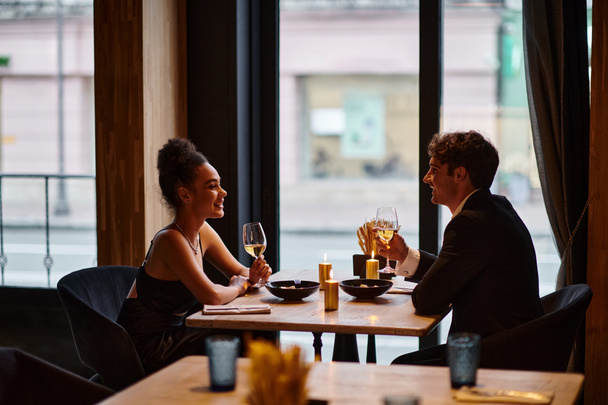happy interracial couple in elegant attire holding glasses with wine during date in restaurant - Photo, Image