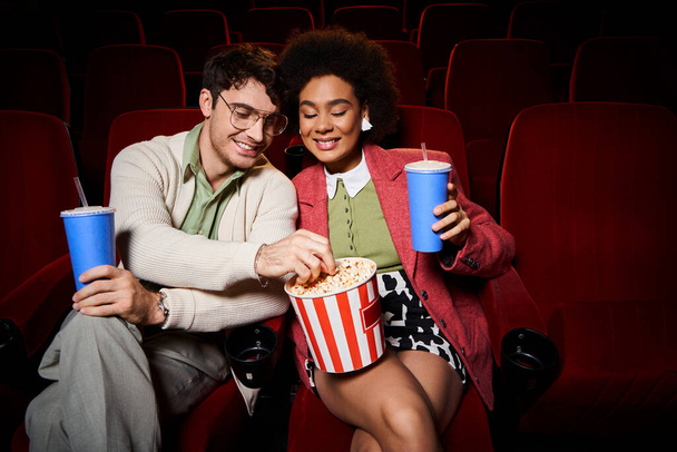 dobrze wyglądająca wesoła, zróżnicowana para w stylowych strojach dzieląca się popcornem w kinie, Walentynki - Zdjęcie, obraz
