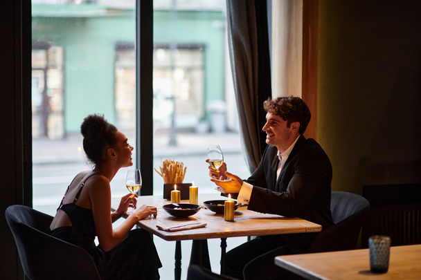 χαρούμενο διαφυλετικό ζευγάρι σε κομψή ενδυμασία κρατώντας ποτήρια με κρασί κατά τη διάρκεια της ημερομηνίας στο εστιατόριο - Φωτογραφία, εικόνα