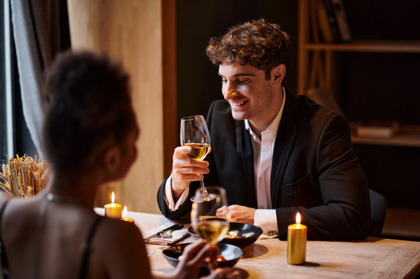 Şık giyinmiş neşeli bir adam restoranda bir kadının yanında şarap kadehine bakıyor. - Fotoğraf, Görsel