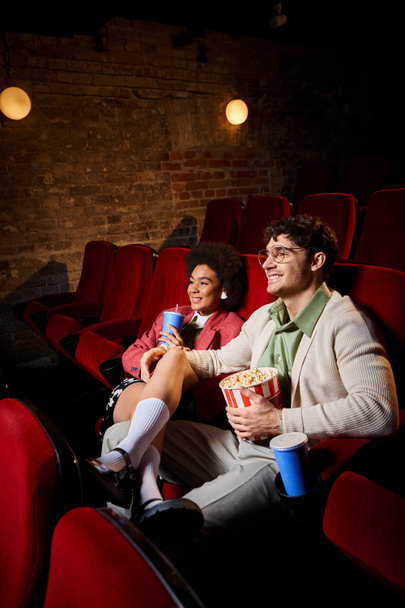 χαρούμενο ποικίλο κομψό ζευγάρι σε ρετρό φορέματα χαμογελώντας και βλέποντας ταινίες σε ημερομηνία στον κινηματογράφο - Φωτογραφία, εικόνα