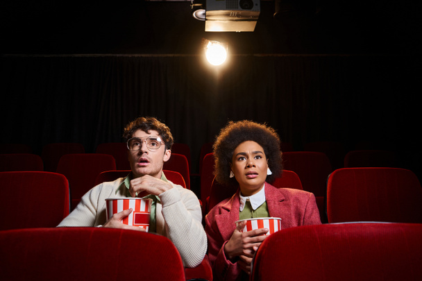 изумленная мультикультурная пара смотрит кино в кино и выглядит удивленно, День Святого Валентина - Фото, изображение