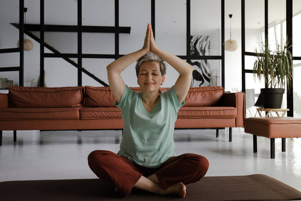 Kobieta w średnim wieku ćwiczy jogę w dużej sali. Uśmiechnięta kobieta siedzi na macie na podłodze w pozycji lotosu i pokazuje namaste gesty dłońmi, uniesionymi nad głową. Spójrz na... - Zdjęcie, obraz