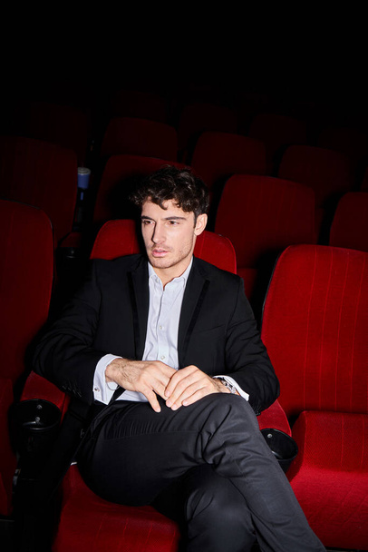 buen aspecto joven modelo masculino con estilo elegante posando en la silla de cine rojo y mirando hacia otro lado - Foto, Imagen