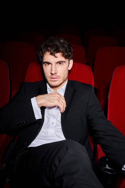 good looking elegant male model in black chic suit sitting on red cinema chair and looking at camera - Photo, Image