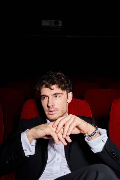 knappe jonge man in zwart elegant pak kijken film in de bioscoop op rode stoelen, handen in de buurt van gezicht - Foto, afbeelding