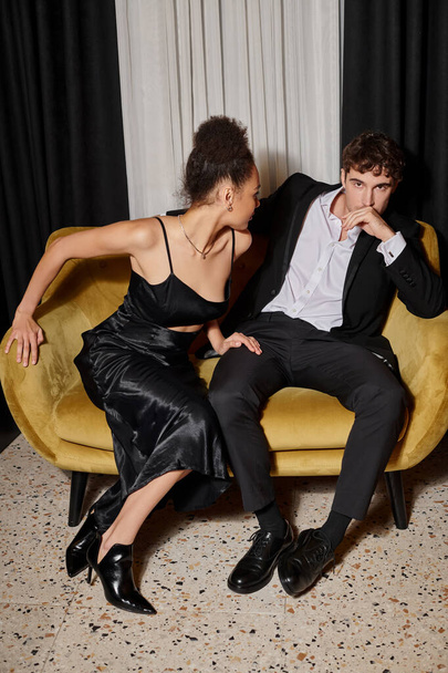 african american woman in black dress looking at handsome man in suit and sitting on velvet sofa - Photo, Image